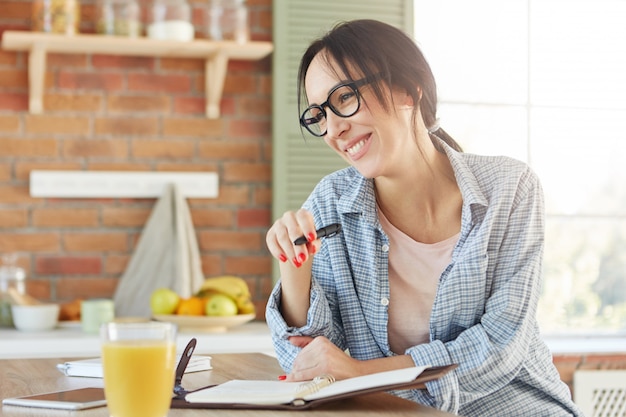 Matin occupé de femme d'affaires. Joyeuse femme souriante exprime des émotions positives comme écrit son horaire de travail dans le journal en spirale,