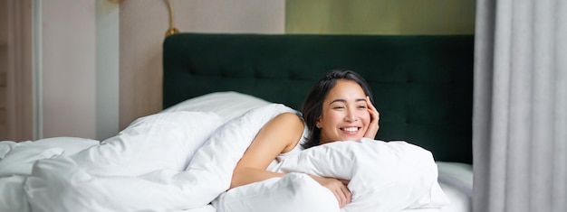 Photo gratuite matins heureux, belle femme coréenne allongée dans son lit souriante, se réveillant optimiste, se couvre de chaleur