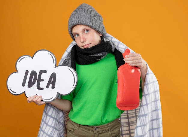 Mauvais petit garçon portant un chapeau chaud et une écharpe enveloppée dans une couverture montrant le signe de la bulle de dialogue avec l'idée de mot et une bouteille d'eau pour garder la chaleur mieux en souriant