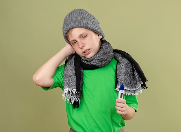 Mauvais petit garçon portant un t-shirt vert en écharpe chaude et un chapeau se sentant mal tenant un thermomètre regardant la caméra malheureux et malade debout sur fond clair