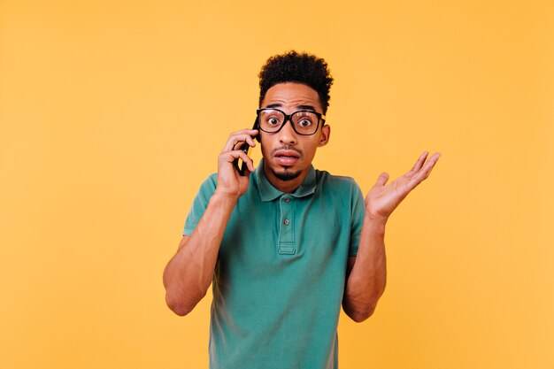 Mec africain choqué dans de grandes lunettes, parler au téléphone. Portrait d'homme bouclé émotionnel posant avec smartphone.