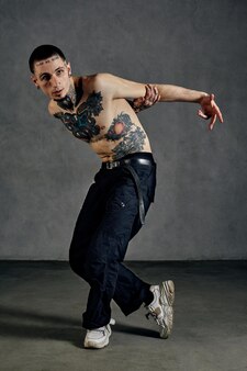 Mec athlétique avec corps et visage tatoués, torse nu, barbe. vêtu d'un pantalon noir et de baskets blanches. il danse sur fond gris studio. dancehall, hip-hop. pleine longueur, espace de copie