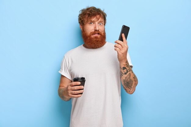 Un mec aux cheveux roux barbu confus tient un téléphone portable, reçoit un appel d'une personne inconnue, entend un cri terrible et fort via un cellulaire, boit du café à emporter, porte un t-shirt blanc décontracté