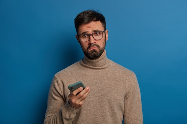 Un mec barbu mécontent sourit, utilise un téléphone portable moderne, a une expression triste, porte des lunettes transparentes et un pull