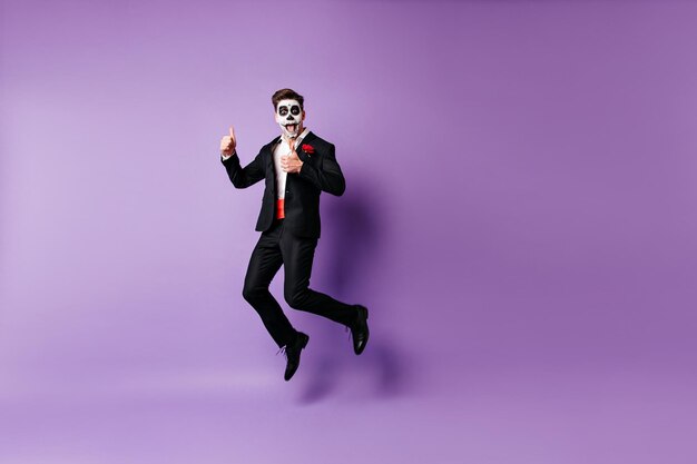 Un mec bien habillé excité avec un maquillage effrayant qui s'amuse en studio Modèle zombie drôle sautant sur fond violet