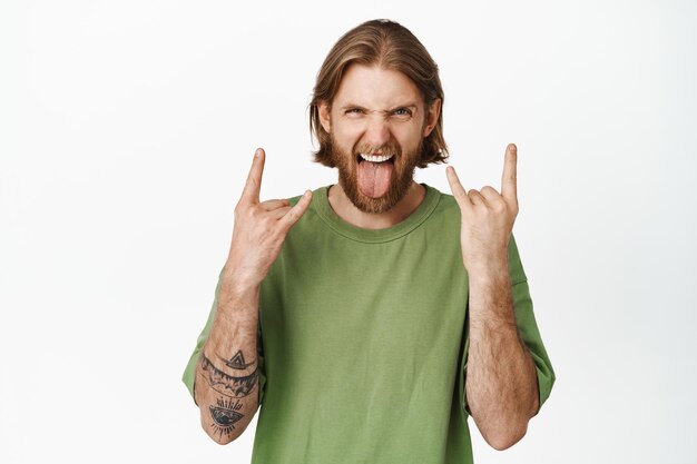 Un mec blond joyeux crie, montre la langue et le signe du heavy metal, rock n roll, profite d'un concert de musique, s'amuse, debout en t-shirt vert sur fond blanc.