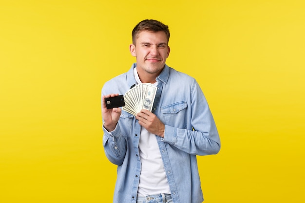 Un mec blond joyeux et ravi qui a l'air excité, souriant satisfait de recevoir un paiement en espèces supplémentaire, tenant une carte de crédit avec de l'argent, debout sur fond jaune optimiste.