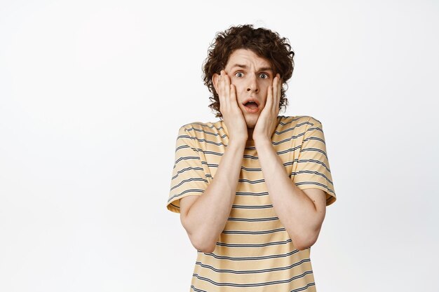 Un mec bouclé effrayé tremblant de peur se tient la main sur le visage et regarde terrifié la caméra debout en t-shirt sur fond blanc