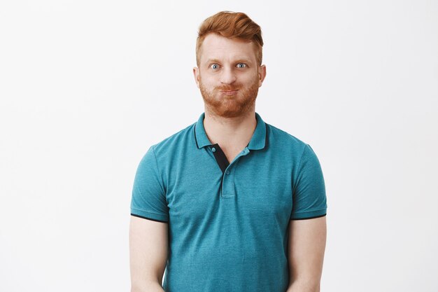 Un mec caucasien mature rousse ludique et insouciant amical avec des poils retenant le souffle à l'intérieur de la bouche et faisant la moue avec une expression et un sourire enfantins, s'amusant sur un mur gris