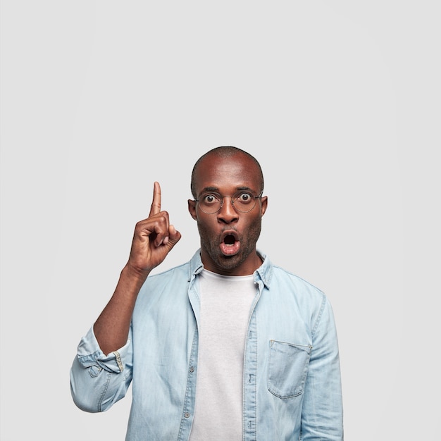 Photo gratuite un mec choqué à la peau foncée regarde avec une expression stupéfaite, pointe son doigt vers le haut, vêtu d'une chemise en jean, se tient contre un mur blanc, remarque quelque chose d'étrange. ethnicité et émotions