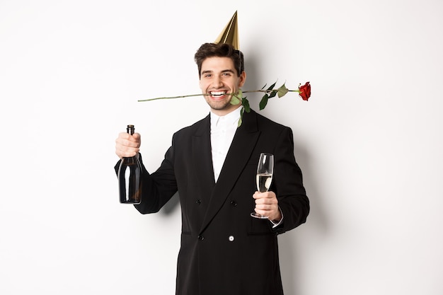 Mec drôle en costume à la mode, célébrant et organisant une fête, tenant une rose dans les dents et du champagne, debout sur fond blanc.