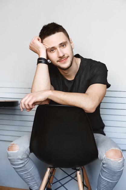 Mec élégant en jeans déchirés, tshirt noir assis sur une chaise.