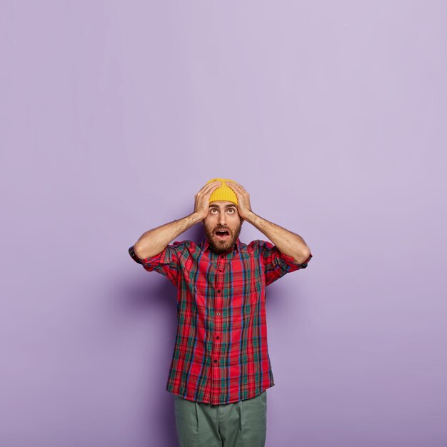 Photo gratuite un mec émotif surpris garde les deux mains sur la tête, porte un chapeau jaune et une chemise à carreaux, a expié le regard vers le haut