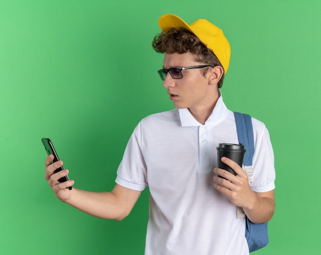 Mec étudiant en chemise blanche et casquette jaune portant des lunettes avec sac à dos tenant un smartphone et une tasse en papier à la confusion et au mécontentement
