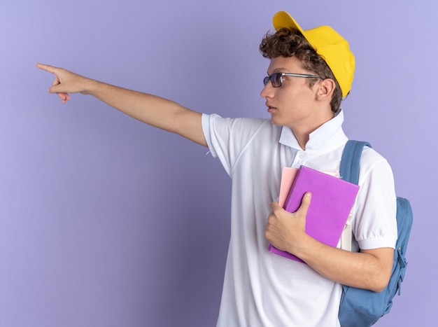 Mec étudiant en polo blanc et casquette jaune portant des lunettes avec sac à dos tenant des cahiers regardant de côté pointant avec l'index quelque chose d'être surpris