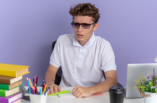 Mec étudiant en polo blanc portant des lunettes assis à la table avec des livres regardant la caméra confus et mécontent sur fond bleu