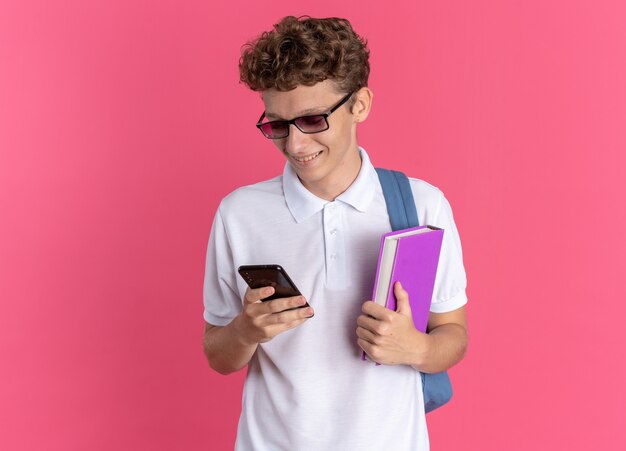 Mec étudiant en vêtements décontractés portant des lunettes avec sac à dos tenant un ordinateur portable et un smartphone regardant l'écran souriant joyeusement debout sur fond rose