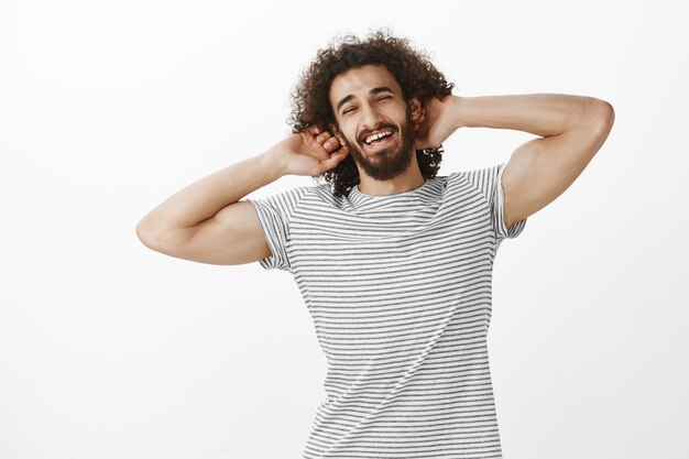 Mec hispanique confiant musculin avec une attitude joyeuse et des cheveux bouclés, tenant les mains derrière la tête et souriant avec une expression insouciante