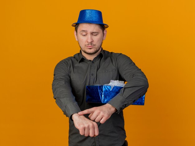 Un mec jeune parti malheureux portant un chapeau bleu tenant une boîte-cadeau et montrant le geste de l'horloge du poignet isolé sur orange