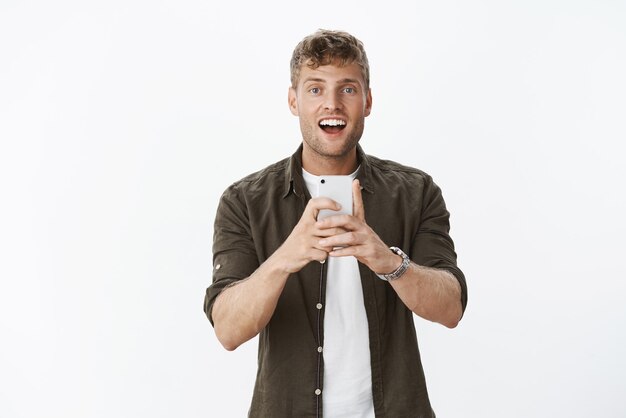 Un mec joyeux et mignon qui dit sourire en prenant une photo sur un smartphone souriant regardant joyeusement la caméra tenant un téléphone portable photographiant sur un téléphone portable avec bonheur et insouciance sur fond gris