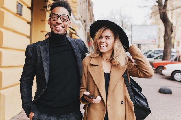 Mec mulâtre souriant en chemise noire posant avec sa charmante amie européenne. Portrait en plein air de fille blonde en riant avec téléphone debout près de jeune homme africain.