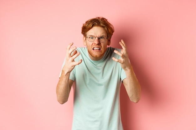 Mec rousse en colère dans des verres en criant, en fronçant les sourcils et en serrant la main avec un visage frustré et indigné, debout sur fond rose.