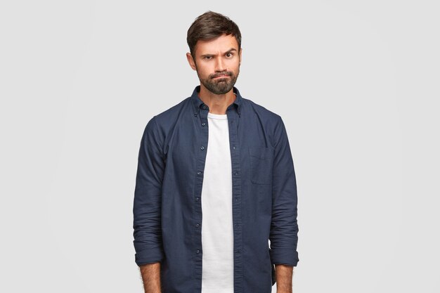 Un mec sombre et mal rasé a une expression irritée, lève les sourcils et serre les lèvres, n'aime pas quelque chose, exprime son mécontentement, porte un t-shirt blanc et une chemise bleu foncé, pose seul