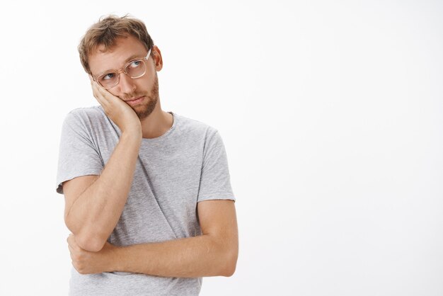 mec sombre nostalgique ennuyé dans des lunettes transparentes avec des poils penchés la tête sur la paume regardant dans le coin supérieur droit avec jalousie et tristesse se sentir ennuyé sur un mur blanc