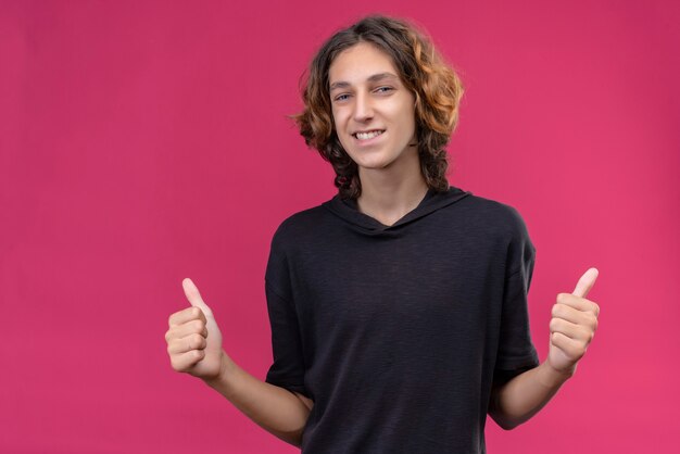 Mec souriant aux cheveux longs en t-shirt noir montrant le pouce vers le haut des deux mains sur le mur rose