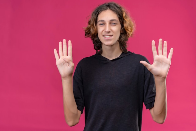 Mec souriant aux cheveux longs en t-shirt noir montrant quatre avec une main et cinq avec l'autre main sur le mur rose