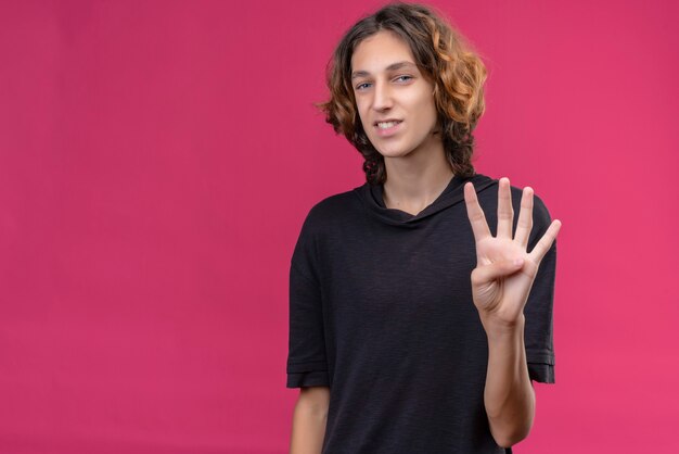 Mec souriant aux cheveux longs en t-shirt noir montrant quatre avec ses doigts sur le mur rose