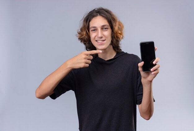 Mec souriant aux cheveux longs en t-shirt noir tenant un téléphone et pointer vers le téléphone sur un mur blanc