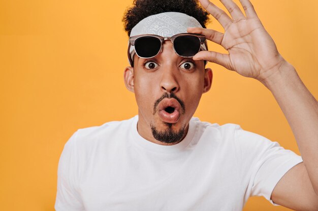 Un mec surpris en tenue blanche enlève ses lunettes et regarde dans la caméra Portrait d'un homme choqué en bandeau posant sur fond orange