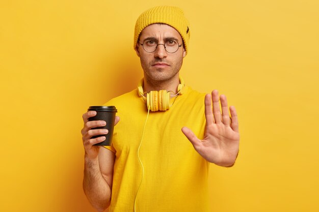 Un mec très mécontent fait un geste d'arrêt, refuse de faire quelque chose, dit non, tient une tasse de café à emporter, porte des lunettes, des vêtements décontractés jaunes