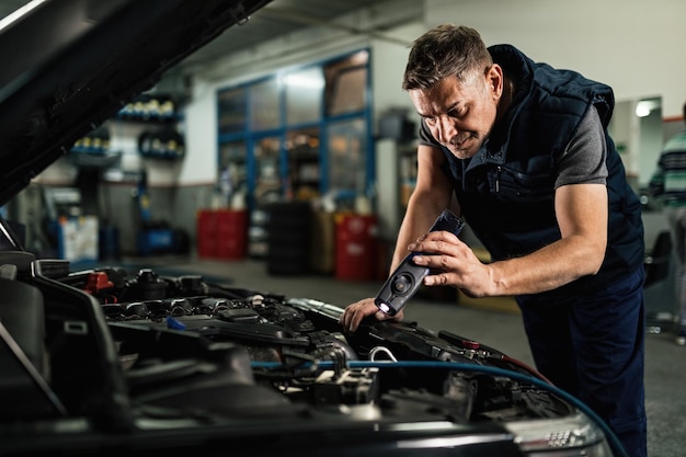 Mécanicien d'âge moyen examinant un moteur de voiture tout en utilisant une lampe dans un atelier de réparation automobile