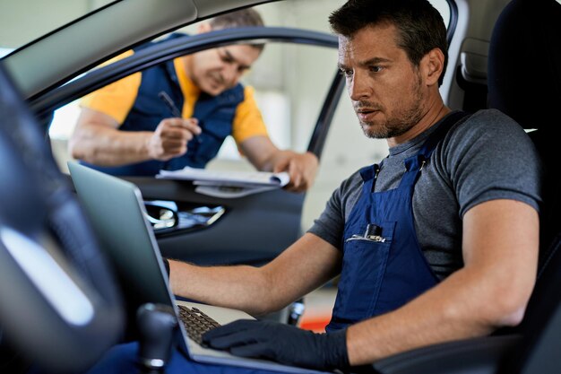 Mécanicien automobile effectuant un diagnostic de voiture sur un ordinateur portable tout en travaillant avec un collègue dans un atelier de réparation automobile