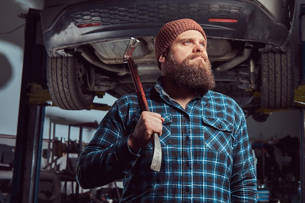 Mécanicien expert barbu vêtu d'une chemise polaire et d'un bonnet, debout avec un pied de biche à la main, contre une voiture sur un ascenseur dans le garage.