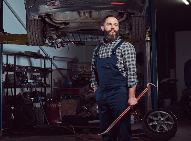 Mécanicien expert barbu vêtu d'un uniforme, debout avec un pied de biche à la main, contre une voiture sur un ascenseur dans le garage.