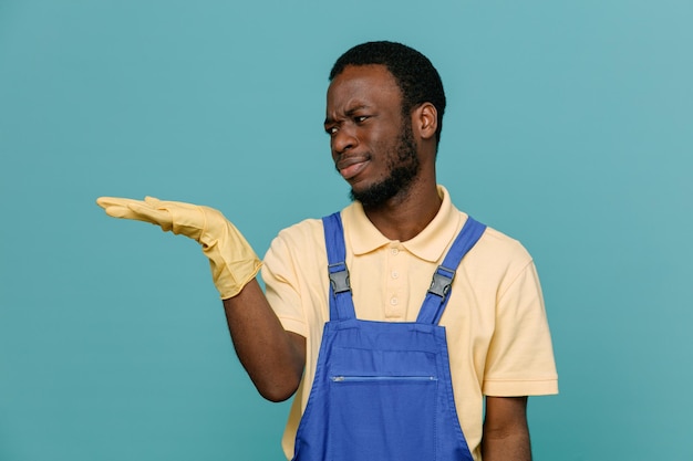 Mécontent de faire semblant de tenir quelque chose de jeune homme nettoyant afro-américain en uniforme avec des gants isolés sur fond bleu