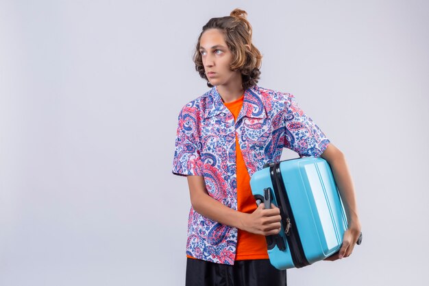 Mécontent jeune beau voyageur guy holding valise sur l'épaule à côté avec une expression triste sur le visage debout