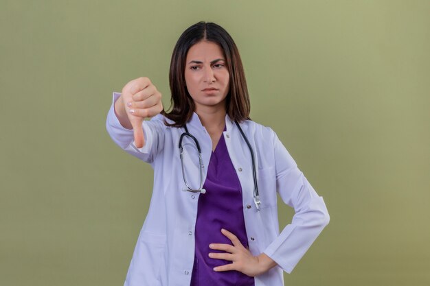 Mécontent jeune femme médecin portant blouse blanche et avec stéthoscope montrant les pouces vers le bas debout sur vert isolé