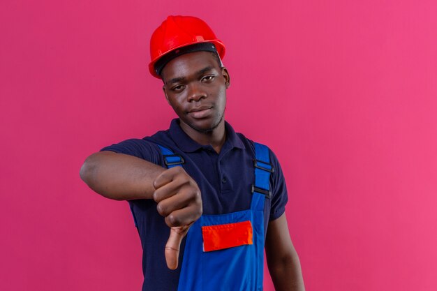 Mécontent jeune homme constructeur afro-américain portant des uniformes de construction et un casque de sécurité montrant les pouces vers le bas insatisfait debout sur rose