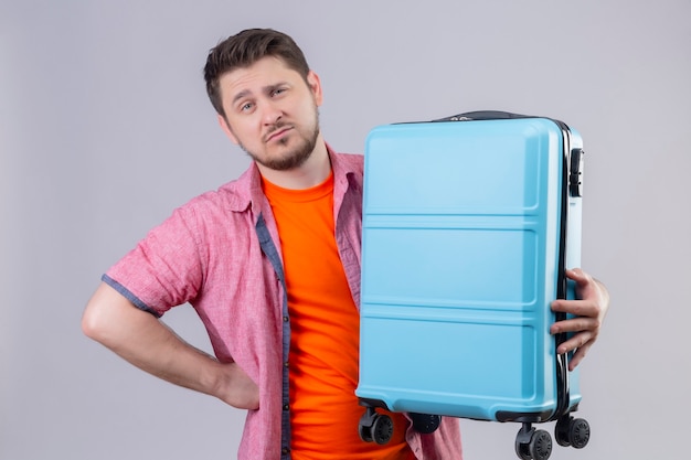 Mécontent jeune voyageur homme tenant une valise bleue avec une expression sceptique sur le visage debout sur un mur blanc