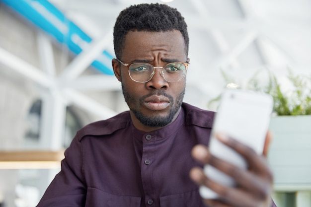 Mécontent malheureux directeur de bureau masculin à la peau sombre regarde avec une expression contrariée au téléphone intelligent, reçoit un message avec de mauvaises nouvelles