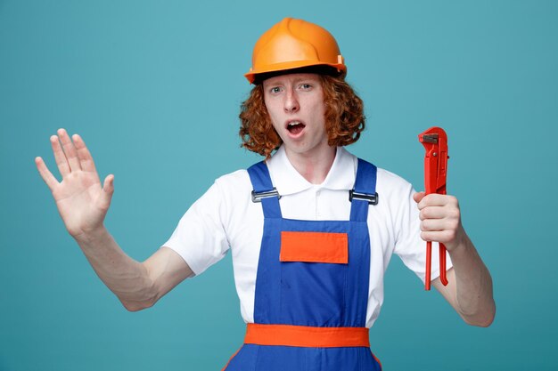 mécontent montrant un geste d'arrêt jeune constructeur homme en uniforme tenant une clé à gaz isolé sur fond bleu