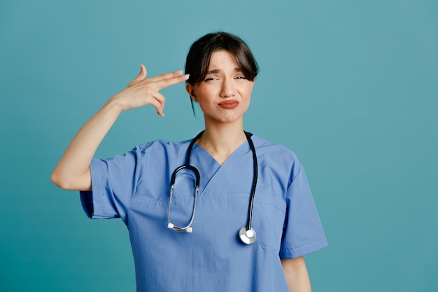 Mécontent montrant un geste de pistolet jeune femme médecin portant un stéthoscope fith uniforme isolé sur fond bleu