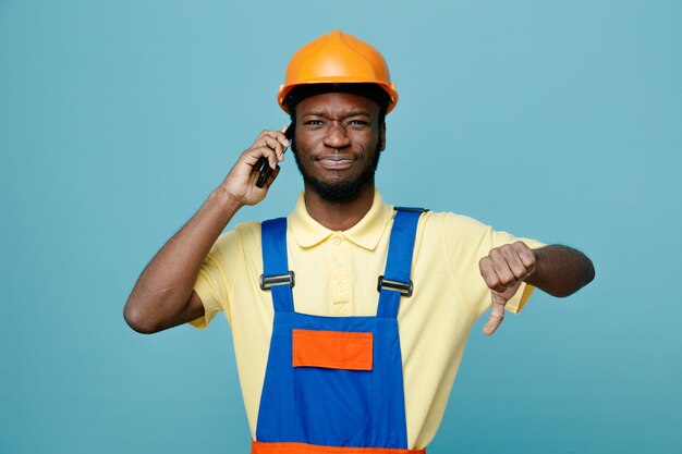 Mécontent montrant les pouces vers le bas jeune constructeur afro-américain en uniforme parle au téléphone isolé sur fond bleu