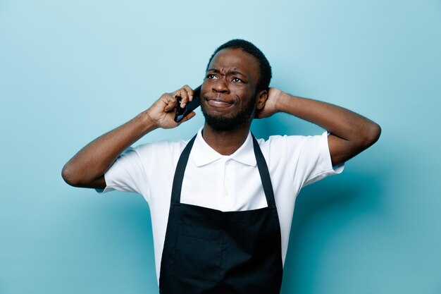 Mécontent parle au téléphone en mettant la main sur la tête jeune coiffeur afro-américain en uniforme isolé sur fond bleu