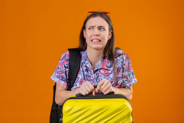 Mécontent et peur jeune voyageur femme portant des lunettes de soleil rouges sur la tête avec sac à dos tenant valise sur mur orange