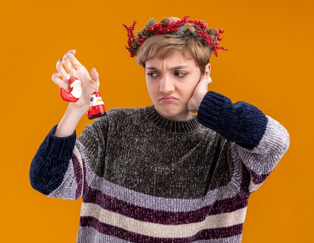 mécontente jeune jolie fille portant une couronne de tête de noël tenant et regardant les ornements de noël du père noël gardant la main sur le visage isolé sur le mur orange